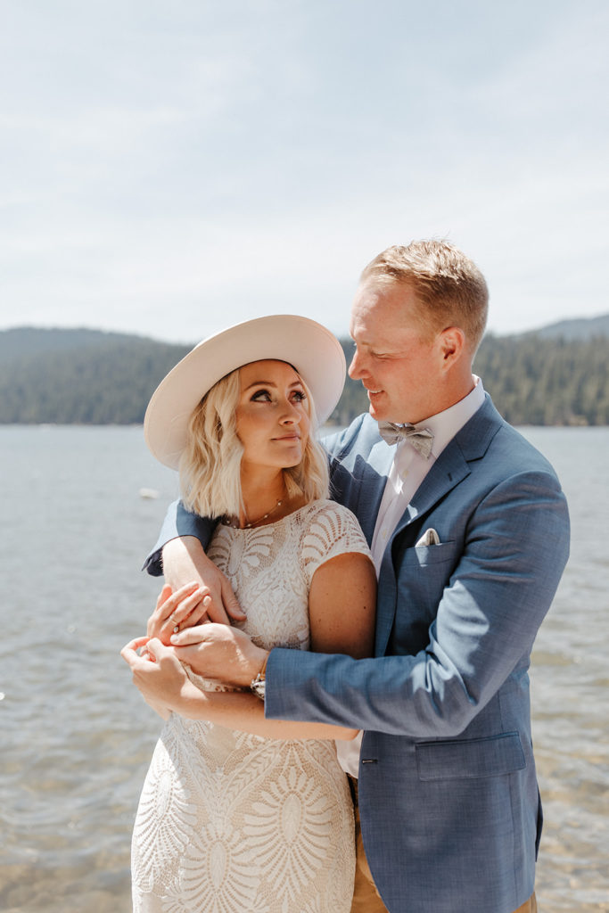 Groom wearing a blue tux wraps his arms around the bride in white with the view of the lake and mountains during their Bend, OR wedding shoot