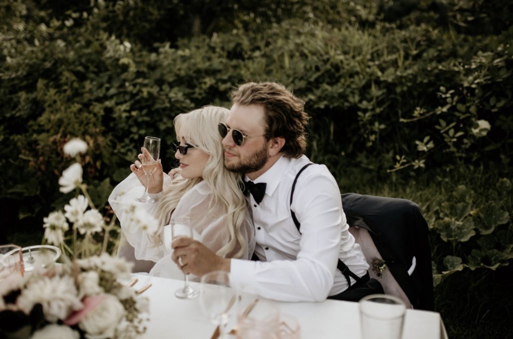 Couple drink a glass of wine and wear sunglasses as they sit on their wedding reception table, captured by Morgan Wirth Photography